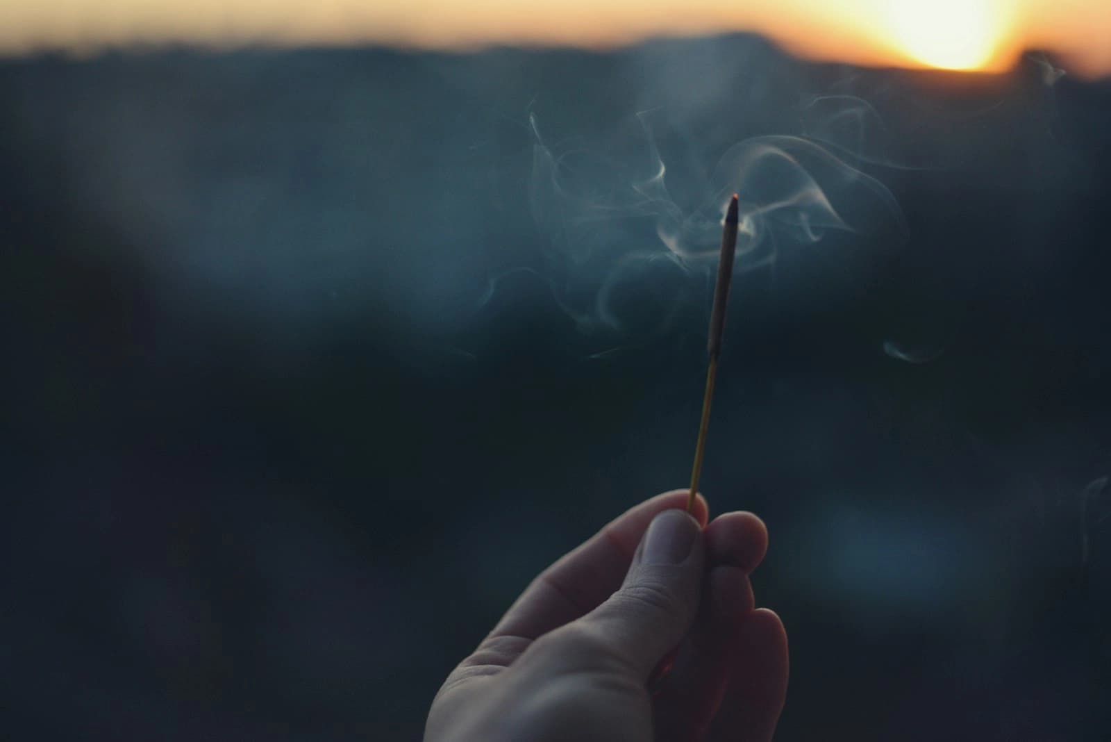 A hand holding a lit incense stick against a dusk sky, smoke curling upward